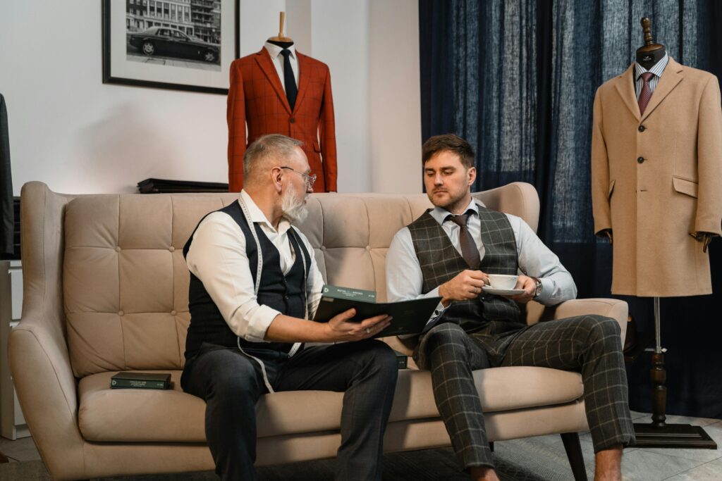 Two men in a tailor shop discussing fashion choices. One holds a book; the other a coffee cup.